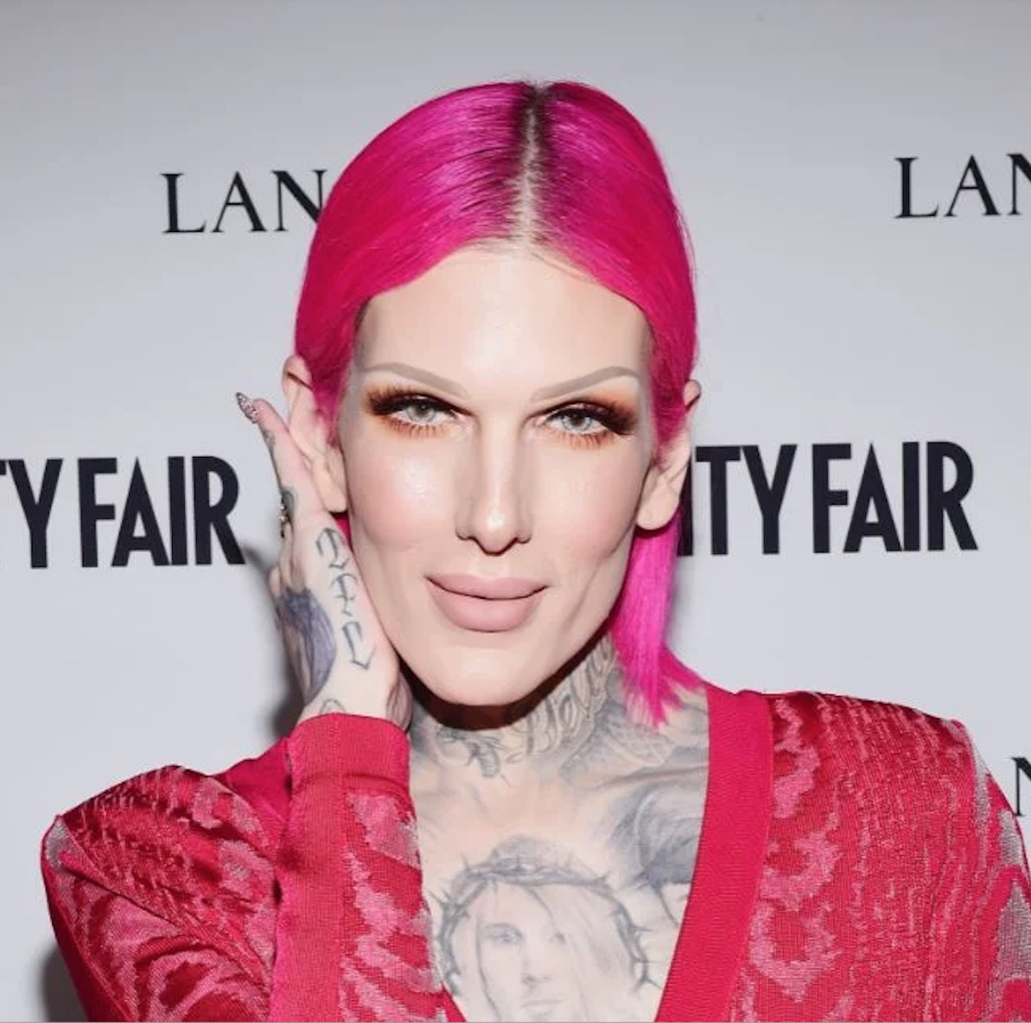 A close-up portrait of Jeffree Star, a fair-skinned person with bright pink hair, posing at a Vanity Fair event. They are wearing a red outfit and have their hand near their ear. The "Vanity Fair" logo is visible in the background.
