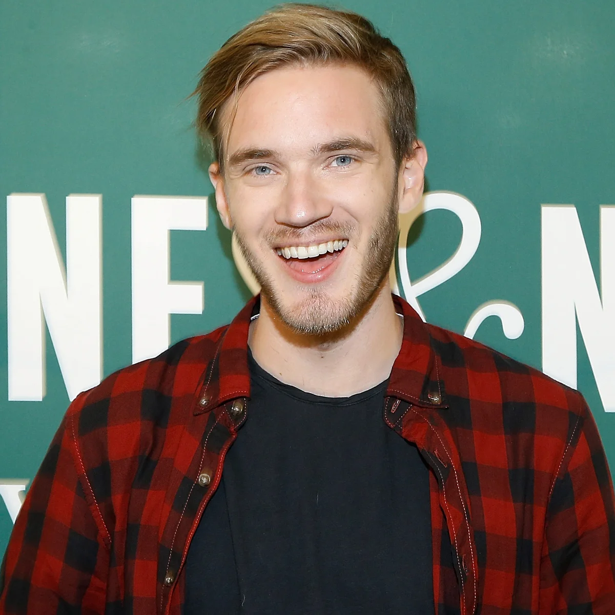 A head-and-shoulders portrait of PewDiePie, a young white man with light brown hair and a short beard, smiling broadly at the camera. He is wearing a red and black flannel shirt over a black t-shirt. The background is a blurred green surface with white letters, possibly a book signing or promotional event.