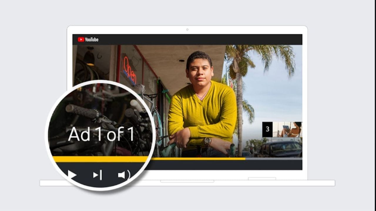 A white laptop screen displays a YouTube video featuring a young man with dark hair wearing a yellow shirt. The video appears to be an advertisement, as indicated by a circle with the text "Ad 1 of 1" and the YouTube logo in the top left corner. A "3" is visible in a smaller thumbnail in the bottom right corner, suggesting this is part of a series or playlist. The laptop is positioned on a gray surface.