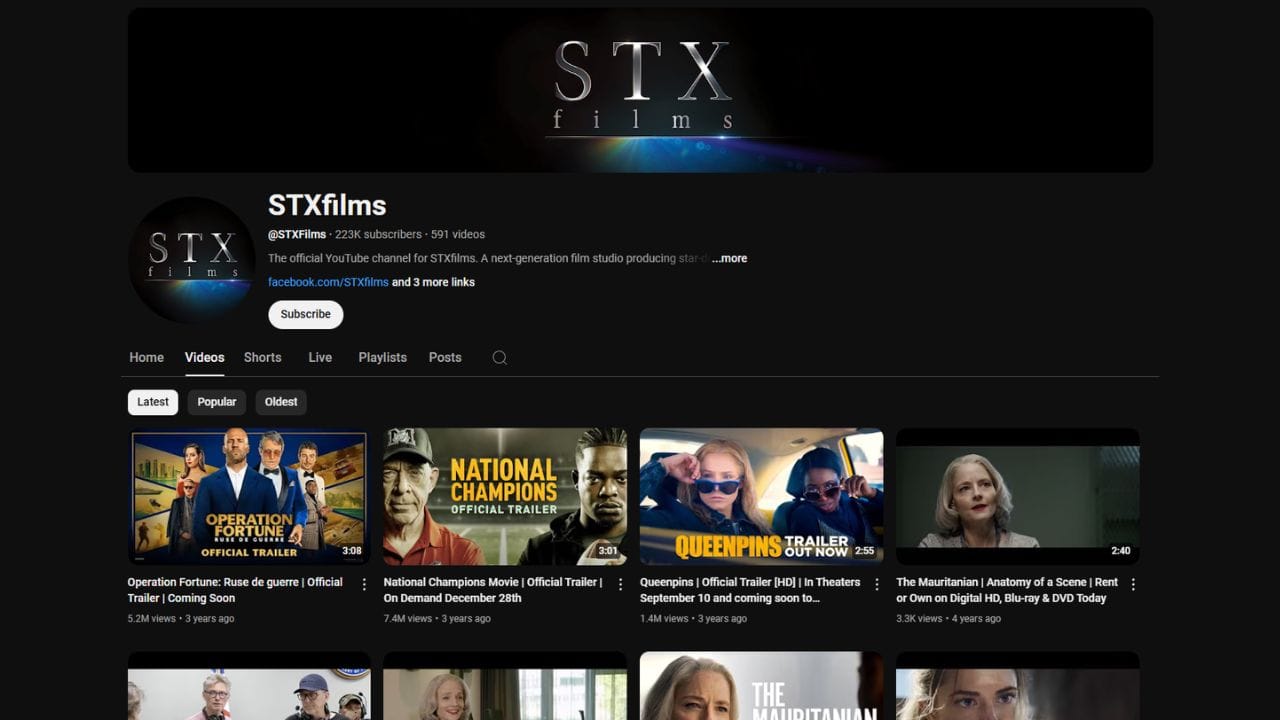 A dark-themed YouTube channel page for STXfilms. The channel banner features the STXfilms logo in white letters on a light gray background. The profile picture is the STXfilms logo on a dark gray background. The channel name "STXfilms" is in white letters, followed by a black dot. Below is the channel username "@STXFilms", the subscriber count (223K), and the video count (591). The banner also includes the text "The official YouTube channel for STXfilms. A next-generation film studio producing star ...more", a link to their Facebook page, and a "Subscribe" button. The channel page displays tabs for "Home," "Videos," "Shorts," "Live," "Playlists," "Posts," and a search icon. Below these tabs are sorting options for "Latest," "Popular," and "Oldest." The video thumbnails show various movie trailers, with titles like "Operation Fortune: Ruse de guerre | Official Trailer | Coming Soon," "National Champions Movie | Official Trailer | On Demand December 28th," "Queenpins | Official Trailer [HD] | In Theaters September 10 and coming soon to...," and "The Mauritanian | Anatomy of a Scene | Rent or Own on Digital HD, Blu-ray & DVD Today."