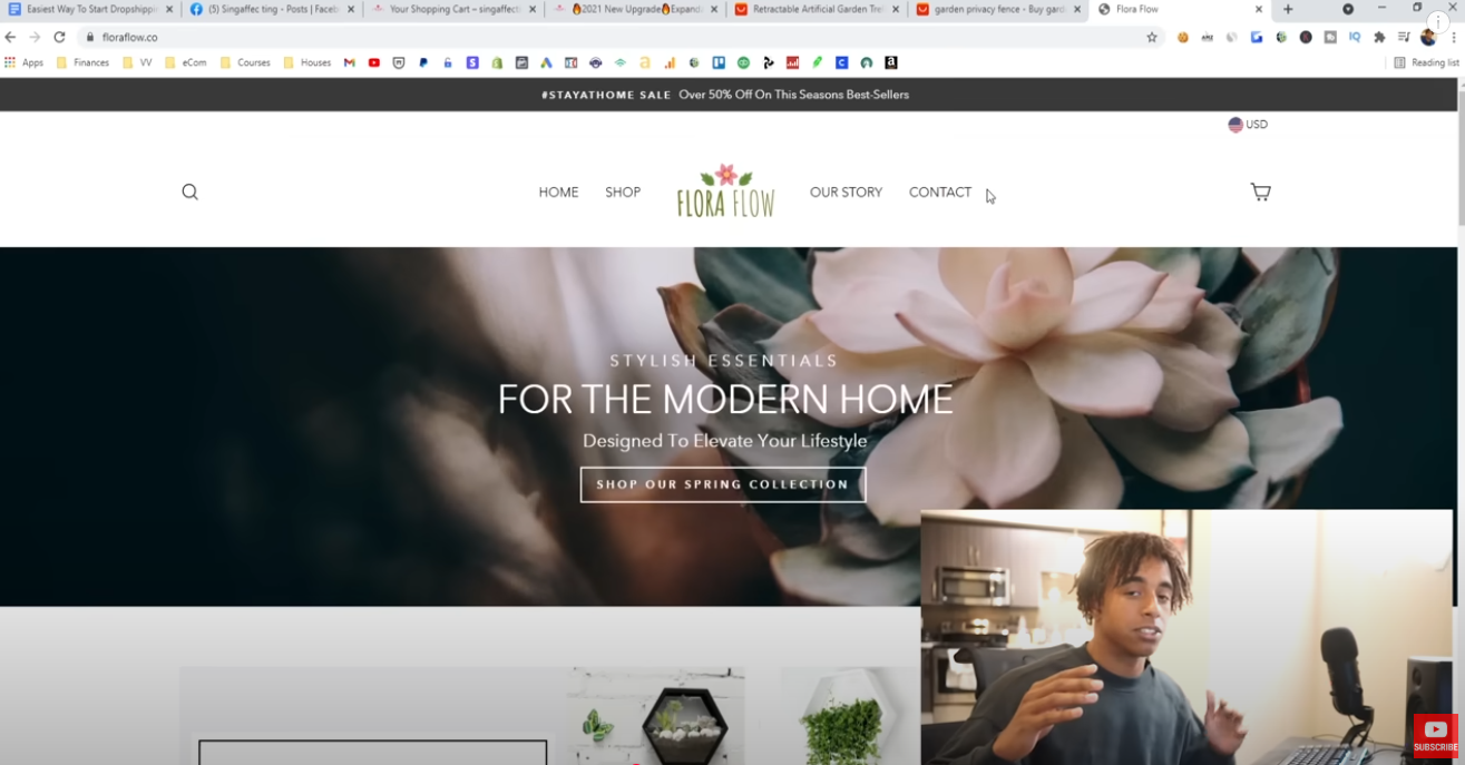 The image is a screenshot of a computer screen. The majority of the screen displays the homepage of the Flora & Flow website. The website features a banner at the top with text about a STAYATHOME SALE offering over 50% off on season's best sellers. The website's logo, "FLORA FLOW" with a floral graphic, is centered in the navigation bar, flanked by "HOME," "SHOP," "OUR STORY," and "CONTACT" links. A search icon and a shopping cart icon are also visible. The main visual on the homepage is a blurred image of light pink flowers with the text "STYLISH ESSENTIALS FOR THE MODERN HOME Designed To Elevate Your Lifestyle SHOP OUR SPRING COLLECTION" overlaid. In the bottom right corner of the screen, a smaller video feed shows Jordan Welch sitting in front of a microphone, gesturing with his hands as if he is speaking or presenting something. A red "SUBSCRIBE" button is visible within the video feed. The overall impression is that Jordan Welch is creating content, possibly a review or walkthrough, related to the Flora & Flow website.