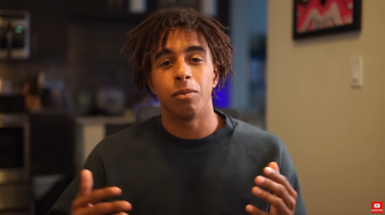 A medium shot of Jordan Welch, a young man with light brown skin and brown dreadlocks. He is wearing a dark gray sweatshirt and looking directly at the camera with a serious but approachable expression. His hands are slightly raised and gesturing as if he is explaining something. The background is slightly blurred, showing a dimly lit room with a dark countertop or shelf to the left and a light beige wall with a framed picture and a light switch to the right. A small red "SUBSCRIBE" button with a white play icon is visible in the bottom right corner, indicating this is likely a frame from a YouTube video. The lighting is soft and even, focusing attention on Jordan Welch.