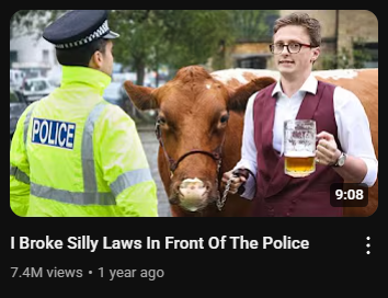 A YouTube video thumbnail featuring three main subjects. On the left, a police officer in a bright yellow jacket with "POLICE" printed on the back and a traditional police hat is facing away from the viewer, looking towards the center. In the center is a brown cow with a white patch on its face, wearing a halter. On the right stands Max Fosh, a young man with glasses, wearing a white shirt, a maroon vest, and holding a large, frothy glass of beer. He is looking towards the camera with a slight smile. The background is blurry but appears to be an outdoor urban setting with cars and buildings. The video duration "9:08" is visible in the bottom right corner of the thumbnail. Below the thumbnail, the video title "I Broke Silly Laws In Front Of The Police" is in white letters. Below the title are the view count "7.4M views" and the upload date "1 year ago." The image suggests a humorous and potentially provocative video where Max Fosh interacts with the police in an unconventional way, possibly involving the cow and the beer.