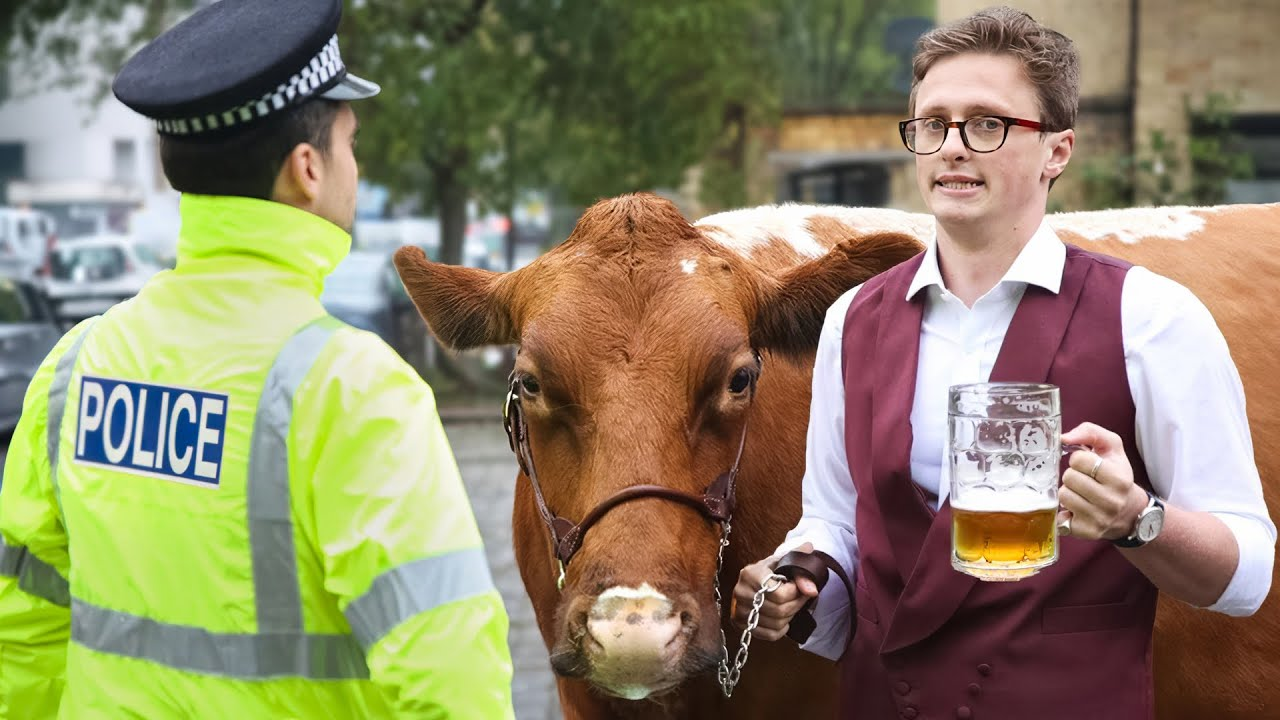 A medium shot of three figures against a blurry urban background. On the left, a police officer in a bright yellow fluorescent jacket with "POLICE" printed in black on the back and a traditional black police cap is facing slightly to the right, looking towards the center of the frame. In the center is a brown Ayrshire cow with white markings on its face and sides, wearing a brown leather halter. Max Fosh, a young man with short brown hair and red-framed glasses, stands on the right, holding a large, frothy glass of beer in his right hand and the cow's lead rope in his left. He is wearing a white collared shirt, a maroon vest, and a watch on his left wrist, and he is looking towards the camera with a slight smile. The background is out of focus but shows parked cars and buildings, suggesting a street scene. The lighting is natural and bright, illuminating the subjects clearly. The overall scene has a slightly comedic and unusual feel, likely a still from one of Max Fosh's videos.